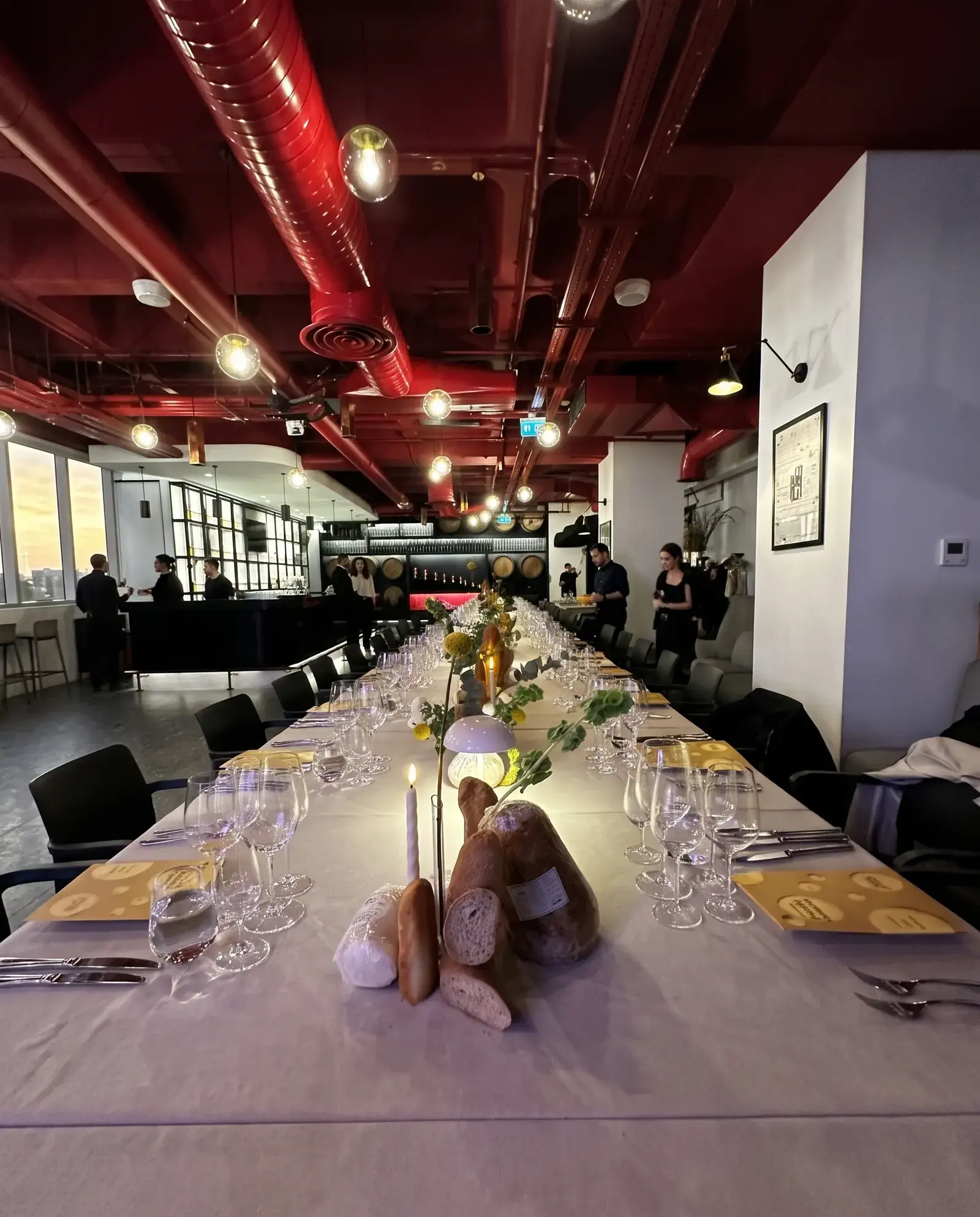 A long dinner table set with bread arrangements, candles and glassware, the bar beyond